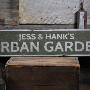 May include: A green wooden sign with white text that reads "Jess & Hank's Urban Garden". The sign is hanging on a wooden wall.
