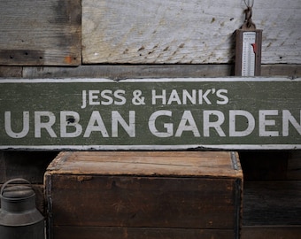 Puede incluir: Un letrero de madera verde con texto blanco que dice "Jess & Hank's Urban Garden". El letrero está colgado en una pared de madera.