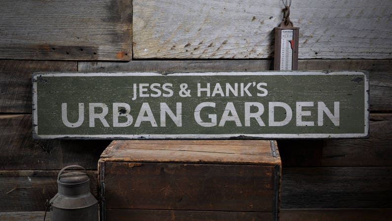May include: A green wooden sign with white text that reads "Jess & Hank's Urban Garden". The sign is hanging on a wooden wall.