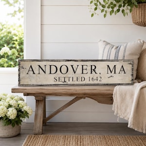 May include: A rectangular wooden sign with a distressed off-white finish and black lettering that reads ANDOVER, MA and SETTLED 1642. The sign features a thin black border and shows signs of wear along the edges, presenting a weathered appearance.
