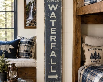 Könnte beinhalten: Ein hohes, rechteckiges, verwittertes dunkelblaues Holzschild mit weißer Aufschrift "BURGESS WATERFALL", gefolgt von einem nach rechts zeigenden Pfeil und dem Text "SCENIC SITE" am unteren Rand. Das Schild hat eine abgenutzte Oberfläche.
