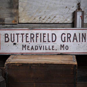 May include: A white wooden sign with red and black checkered squares on each end. The sign reads "Butterfield Grain Meadville, Mo".