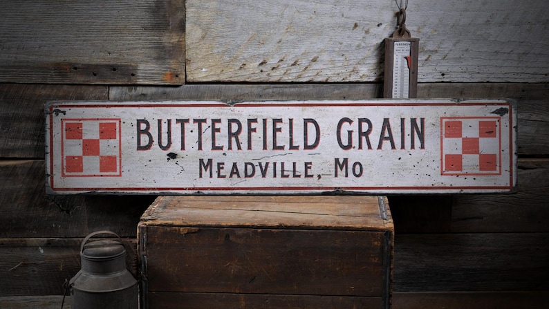 May include: A white wooden sign with red and black checkered squares on each end. The sign reads "Butterfield Grain Meadville, Mo".