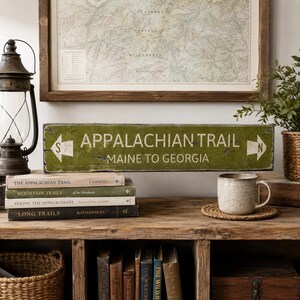 May include: A rectangular green wooden sign with distressed edges featuring white text that reads APPALACHIAN TRAIL and MAINE TO GEORGIA, flanked by two white arrows pointing in opposite directions, labeled with the letters S and N on each side.