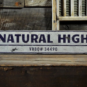 May include: A rectangular metal sign with a distressed white finish and dark blue border. The text "NATURAL HIGH" is printed in large dark blue capital letters, with "VRBO# 34490" printed in smaller dark blue text centered below the main line.