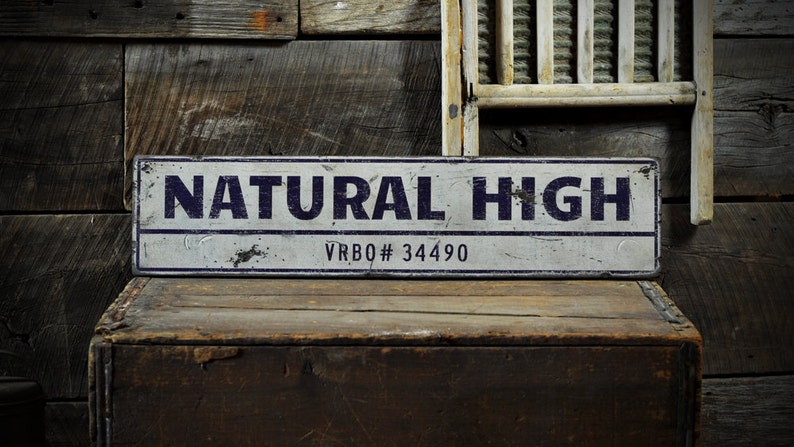 May include: A rectangular metal sign with a distressed white finish and dark blue border. The text "NATURAL HIGH" is printed in large dark blue capital letters, with "VRBO# 34490" printed in smaller dark blue text centered below the main line.
