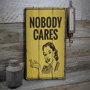 May include: A yellow wooden sign with black text that reads "Nobody Cares". A woman with a surprised expression is pictured on the sign. The sign is hanging on a rustic wooden wall.