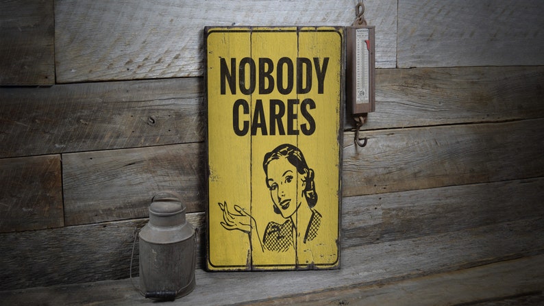 May include: A yellow wooden sign with black text that reads "Nobody Cares". A woman with a surprised expression is pictured on the sign. The sign is hanging on a rustic wooden wall.