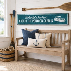 Op de afbeelding: Een rechthoekig blauwgroen wandbord met witte tekst Nobody's Perfect EXCEPT THE PONTOON CAPTAIN en een witte afbeelding van een pontonboot. Het bord is bevestigd aan een witte wand van schrootjes boven een houten bank met drie kussens.