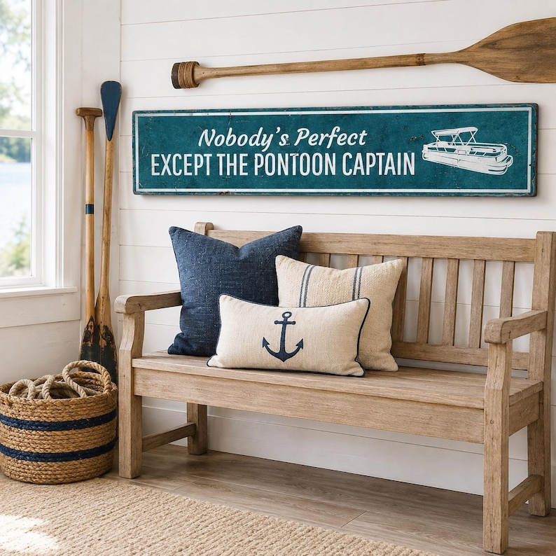 Op de afbeelding: Een rechthoekig blauwgroen wandbord met witte tekst Nobody's Perfect EXCEPT THE PONTOON CAPTAIN en een witte afbeelding van een pontonboot. Het bord is bevestigd aan een witte wand van schrootjes boven een houten bank met drie kussens.