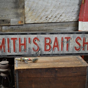 Könnte beinhalten: Ein verwittertes Schild mit der Aufschrift "SMITH'S BAIT SHOP" in roten Buchstaben. Das Schild ist an einer rustikalen Holzwand angebracht. Eine Holzkiste, ein Angelköder und eine alte Laterne im Vordergrund, passend zum Thema Angeln.