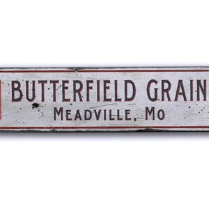 May include: A white wooden sign with red and black lettering that reads "Butterfield Grain Meadville, MO". The sign has two red and white checkered squares on either side of the text.