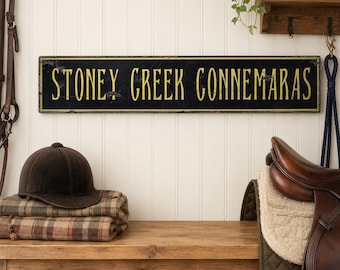 May include: A rectangular black wooden sign with a thin gold border and the yellow text STONEY CREEK CONNEMARAS in a serif font. The sign is mounted on a white paneled wall above a wooden bench with a brown riding helmet and saddle.