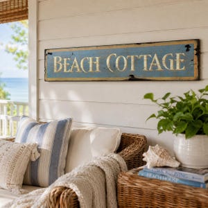May include: A rectangular horizontal wooden sign with a distressed blue finish and gold-colored lettering that reads BEACH COTTAGE. The sign features a weathered texture with visible dark edges and a simple, clean font style.