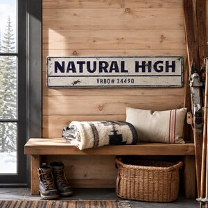 May include: A rectangular white metal sign with a distressed finish featuring the dark blue text NATURAL HIGH and the smaller text VRB0# 34490. The sign has a thin dark blue horizontal line separating the two lines of text.