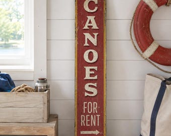 Könnte beinhalten: Ein hohes, rechteckiges Holzschild mit abgenutzter roter Oberfläche und gelben Kanten. Das Schild zeigt weiße Blockbuchstaben mit der Aufschrift CANOES FOR RENT sowie einen Pfeil nach rechts und den Schriftzug Lake Monroe in Schreibschrift.