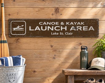 Könnte beinhalten: Ein rechteckiges braunes Schild mit weißer Aufschrift CANOE & KAYAK LAUNCH AREA und Lake St. Clair sowie einem weißen Symbol einer Person im Kajak. Das Schild hat eine abgenutzte Oberfläche und ist an einer hellbraunen Holzwand befestigt.