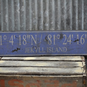 May include: A weathered blue wooden sign with white text. The sign reads "31° 4' 18"N / 81° 24' 16"W JEKYLL ISLAND".