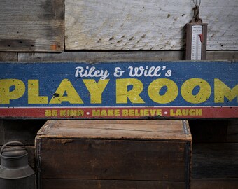 May include: A blue and yellow wooden sign with the text "Riley & Will's Playroom" in white. The sign also has the text "Be Kind - Make Believe - Laugh" in white on a red and white stripe.