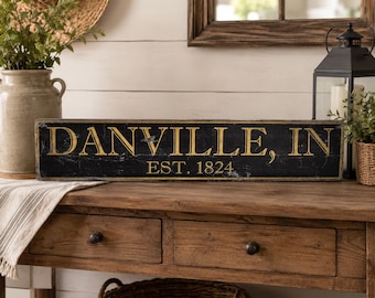 Può includere: Un'insegna rettangolare in legno con finitura nera usurata e scritte dorate che recitano DANVILLE, IN e EST. 1824. L'insegna presenta una texture invecchiata con segni di usura visibili lungo i bordi e sulla superficie, conferendo un aspetto antico.