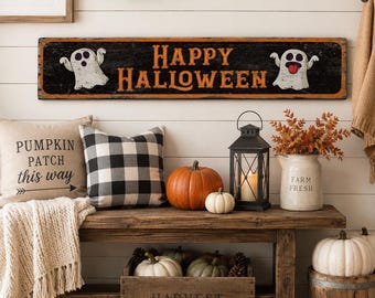 May include: A long rectangular wooden sign with a black background and orange border featuring the text "HAPPY HALLOWEEN" in orange letters, flanked by two white ghost illustrations, one with a tongue sticking out, mounted on a white wall.