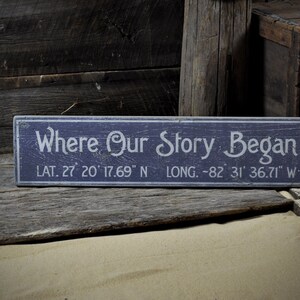 May include: A blue wooden sign with white text that reads "Where Our Story Began" and includes the latitude and longitude coordinates: "LAT. 27 20 17.69" N LONG. -82 31 36.71" W.