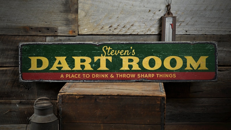May include: A rustic wooden sign with a green background and red trim. The sign reads "Steven's Dart Room" in yellow letters and "A Place to Drink & Throw Sharp Things" in smaller yellow letters.