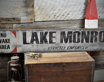 Könnte beinhalten: Ein rustikales Holzschild mit schwarzer Beschriftung, auf dem "LAKE MONROE" und "STRICTLY ENFORCED" steht. Auf der linken Seite des Schildes befindet sich ein roter und weißer Kreis mit dem Text "NO WAKE AREA".