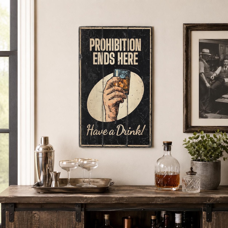 May include: A rectangular wall sign with a black background featuring the text PROHIBITION ENDS HERE and Have a Drink! in white and cream lettering. A central illustration shows a hand holding a glass of amber liquid inside a cream oval.