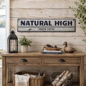 May include: A rectangular metal sign with a distressed white finish and black border. The text "NATURAL HIGH" is printed in large, bold, dark blue capital letters, with "VRBO# 34490" printed in smaller black text on a horizontal line below.