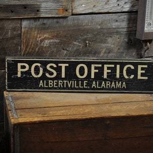 May include: Black wooden sign with white text that reads "POST OFFICE ALBERTIVILLE, ALABAMA". The sign is sitting on a wooden crate.