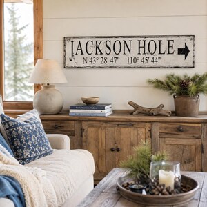 May include: A rectangular wall sign with a distressed white finish and black lettering that reads JACKSON HOLE, followed by the coordinates N 43&deg; 28' 47" 110&deg; 45' 44" and a black arrow pointing to the right side of the sign.