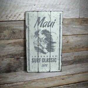 May include: A rustic wooden sign with a faded green background and white lettering. The sign says "Maui Longboard Surf Classic 1976" and features a black and white illustration of a surfer riding a wave.