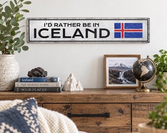 May include: A rectangular white metal sign with a distressed finish featuring the black text I'D RATHER BE IN ICELAND and a graphic of the Icelandic flag on the right side. The sign has a thin black border and is mounted on a plain wall.