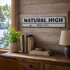 May include: A rectangular metal sign with a distressed white finish and dark blue lettering that reads NATURAL HIGH and VRBO# 34490. The sign is mounted on a horizontal wood plank wall above a desk with books, a plant, and a lamp.