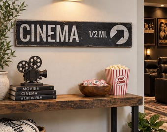 以下が含まれることがあります： 端が擦れた長方形の黒い木製看板。白い文字で「CINEMA 1/2 MI.」と右向きの白い矢印が描かれています。看板は壁に取り付けられており、その下には映画用プロジェクターの模型と本が置かれた木製のコンソールテーブルがあります。