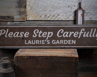 Puede incluir: Un letrero de madera marrón con texto blanco que dice "Please Step Carefully Laurie's Garden". El letrero está colgado en una pared de madera rústica.