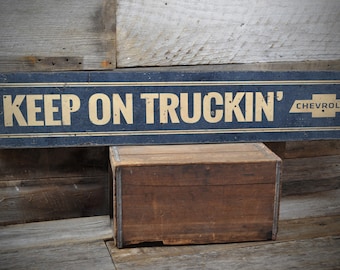 Può includere: Un cartello in legno nero con lettere dorate che recita "KEEP ON TRUCKIN'" con il logo Chevrolet in oro sul lato destro.
