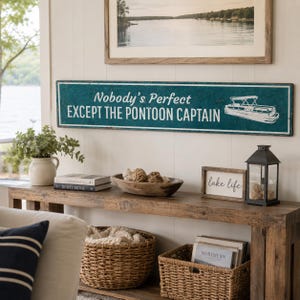 Op de afbeelding: Een lang, rechthoekig metalen bord in een blauwgroene kleur met een verweerde afwerking. Het bord bevat witte tekst met de tekst "Nobody's Perfect EXCEPT THE PONTOON CAPTAIN" en een witte lijntekening van een pontonboot aan de rechterkant.