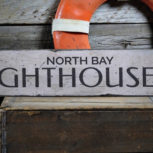 May include: A weathered wooden sign with white paint and black lettering that reads "North Bay Lighthouse" with a black arrow pointing to the right. The sign is hanging on a rustic wooden wall with a red and white life preserver hanging above it.