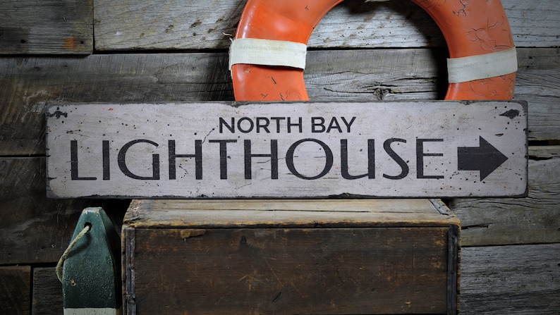 May include: A weathered wooden sign with white paint and black lettering that reads "North Bay Lighthouse" with a black arrow pointing to the right. The sign is hanging on a rustic wooden wall with a red and white life preserver hanging above it.