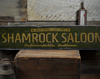 Può includere: Un cartello in legno verde con lettere nere che recita "Shamrock Saloon" con il testo "Indianapolis, Indiana" e "Good Friends, Good Times" in caratteri più piccoli.