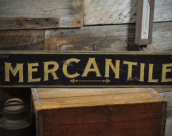 Op de afbeelding: Een rechthoekig zwart bord met het woord "MERCANTILE" in gouden letters. Het bord is licht beschadigd en heeft een decoratief pijldesign. Het bord wordt weergegeven tegen een rustieke houten achtergrond, met een houten kist en een metalen container op de voorgrond.