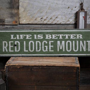 May include: A rustic green wooden sign with white lettering that reads "Life is better on Red Lodge Mountain".
