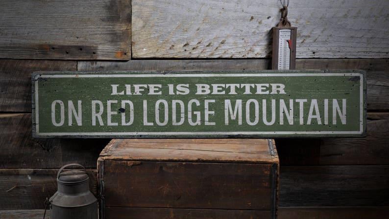 May include: A rustic green wooden sign with white lettering that reads "Life is better on Red Lodge Mountain".