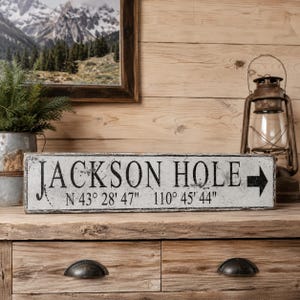 May include: A rectangular wooden sign with a distressed white finish and black lettering that reads JACKSON HOLE, followed by the coordinates N 43&deg; 28' 47" 110&deg; 45' 44" and a black arrow pointing to the right side of the sign.