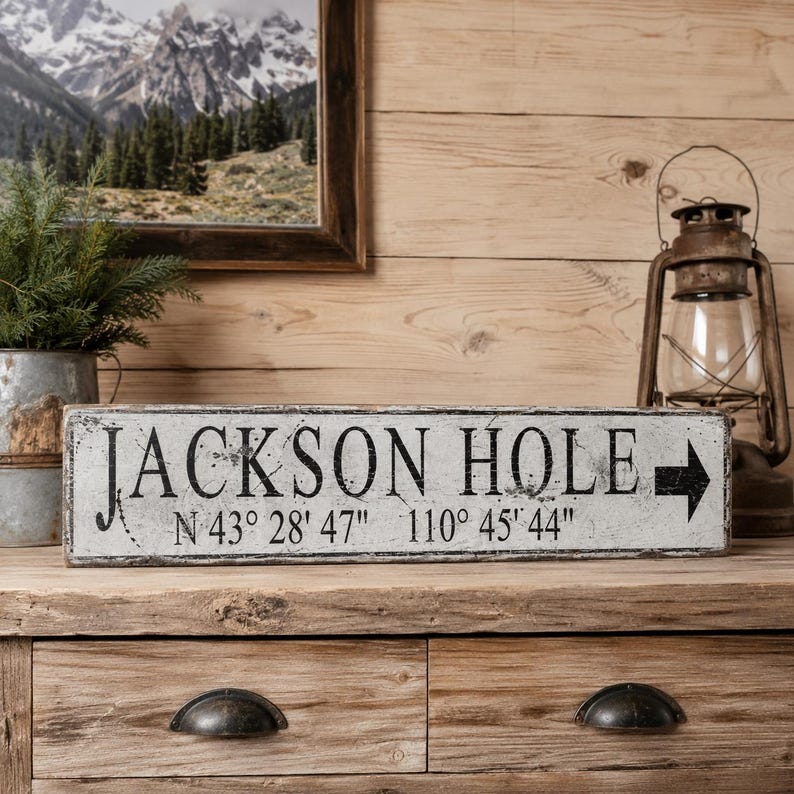 May include: A rectangular wooden sign with a distressed white finish and black lettering that reads JACKSON HOLE, followed by the coordinates N 43&deg; 28' 47" 110&deg; 45' 44" and a black arrow pointing to the right side of the sign.