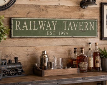 Può includere: Un'insegna da parete rettangolare verde con finitura anticata che riporta la scritta bianca RAILWAY TAVERN e EST. 1994. L'insegna presenta un sottile bordo bianco ed è montata orizzontalmente su una parete in assi di legno chiaro.