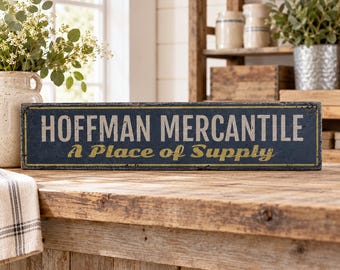 Op de afbeelding: Een rechthoekig houten bord met een donkerblauwe afwerking en versleten randen. De tekst HOFFMAN MERCANTILE is gedrukt in grote, lichtgekleurde hoofdletters, met de zin A Place of Supply in een kleiner, goudkleurig cursief lettertype eronder.