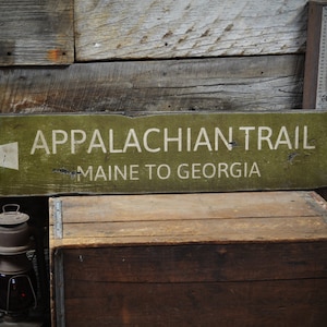 May include: A rustic wooden sign with green paint and white lettering. The sign reads "Appalachian Trail" with arrows pointing north and south. The text below the trail name reads "Maine to Georgia".